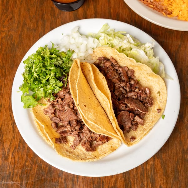 Two carne asada tacos with cilantro, onions, and lettuce on a white plate.