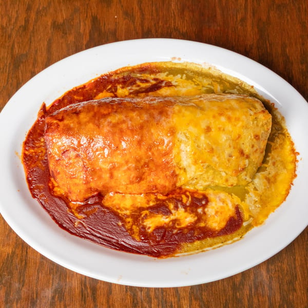 A large, cheese-covered burrito on a white plate, half smothered in red sauce and half in green sauce.