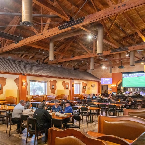 A spacious restaurant interior with high wooden ceilings, patrons dining, and a large TV screen showing sports.