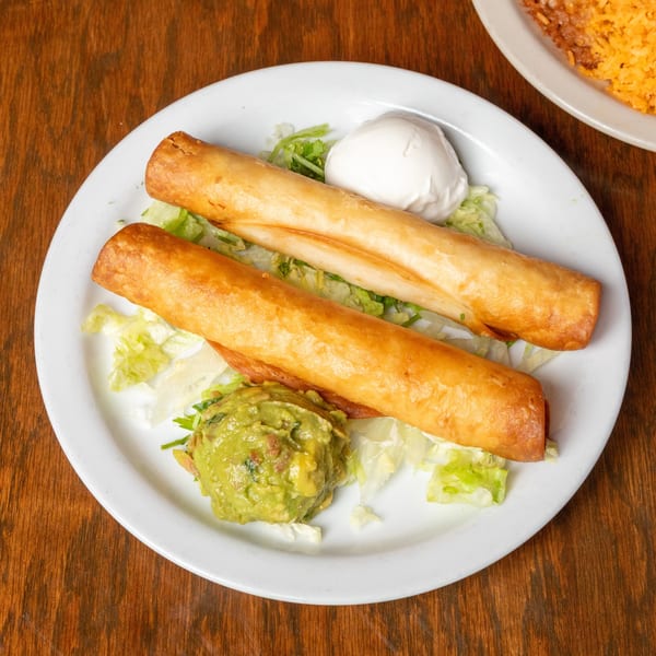Two golden taquitos on a bed of lettuce with guacamole and sour cream on a white plate.